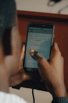 A person is holding a smartphone with both hands, reading a chat conversation on the screen. The chat messages are visible, containing text and emojis. The background is blurred and has a warm tone, indicating a possibly indoor environment.