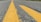 A close-up view of a roadway with a prominent yellow line running down the middle, showing the textured asphalt. The perspective is low to the ground, creating a sense of depth as the road stretches into the distance. Green grass is visible on the sides, indicating a rural or suburban setting.
