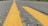 A close-up view of a roadway with a prominent yellow line running down the middle, showing the textured asphalt. The perspective is low to the ground, creating a sense of depth as the road stretches into the distance. Green grass is visible on the sides, indicating a rural or suburban setting.
