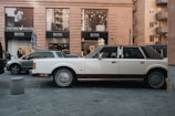 A vintage white and brown car is parked on a street in front of a modern building with large glass windows displaying upscale clothing brands like Emporio Armani and Hugo Boss. The street is busy with other parked vehicles and pedestrians walking by. The building features a neutral stone facade, and various signs are visible near the pavement.