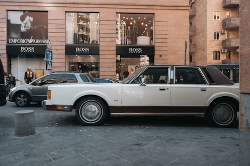 A vintage white and brown car is parked on a street in front of a modern building with large glass windows displaying upscale clothing brands like Emporio Armani and Hugo Boss. The street is busy with other parked vehicles and pedestrians walking by. The building features a neutral stone facade, and various signs are visible near the pavement.