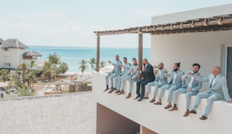 A group of men dressed in light blue suits and one in a dark suit are sitting on a ledge of a building, each holding a drink. They appear relaxed and cheerful, with the backdrop of a tropical seaside location featuring palm trees and ocean.