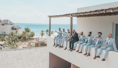 A group of men dressed in light blue suits and one in a dark suit are sitting on a ledge of a building, each holding a drink. They appear relaxed and cheerful, with the backdrop of a tropical seaside location featuring palm trees and ocean.