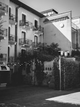 A multi-story building with European architecture featuring balconies with wrought iron railings. The building has a sign reading 'Hotel Catullo' on the facade. Vines climb over parts of the exterior, and string lights hang above a courtyard area. The scene is cast in black and white, providing a nostalgic atmosphere.