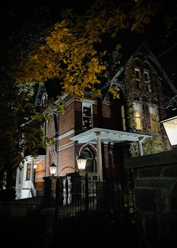 A large, old brick house is depicted at night, partially obscured by trees with yellow leaves. The house features gothic architectural elements such as pointed arches and ornate detailing. Lantern-style lights illuminate the entrance, casting shadows on the surroundings. The structure is surrounded by a metal fence.
