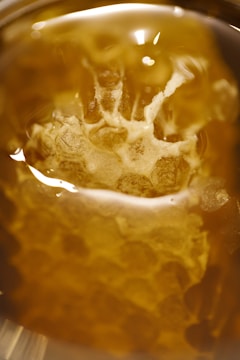 Close-up of a honeycomb structure immersed in golden, translucent liquid. The hexagonal pattern is visible through the liquid, and light reflections add an ethereal glow.