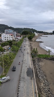 A coastal roadway with several vehicles and motorbikes traveling along it, bordered by a sidewalk and lush trees. To the right, a beach with sand and rocks stretches alongside the ocean, with waves gently lapping the shore. In the background, there are several buildings, including a mosque with prominent domes, set against a backdrop of green hills and a cloudy sky.