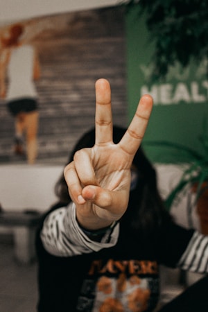 A hand is prominently displaying a peace sign gesture in the foreground. The fingers are clearly in focus, while the background is blurred, showing parts of an indoor setting with a wall, some text, and a plant.