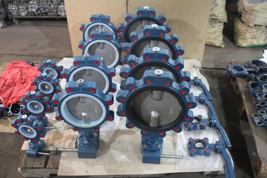 A collection of industrial butterfly valves arranged in rows on a warehouse floor. The valves are primarily metallic with blue and red accents and are placed on top of white packaging material. Additional valves and mechanical components are visible in the background alongside red fabric.