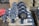 A collection of industrial butterfly valves arranged in rows on a warehouse floor. The valves are primarily metallic with blue and red accents and are placed on top of white packaging material. Additional valves and mechanical components are visible in the background alongside red fabric.