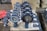 A collection of industrial butterfly valves arranged in rows on a warehouse floor. The valves are primarily metallic with blue and red accents and are placed on top of white packaging material. Additional valves and mechanical components are visible in the background alongside red fabric.