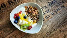 A bowl filled with creamy yogurt topped with a variety of fresh fruits and nuts. Slices of banana and kiwi are arranged alongside blueberries, red currants, and cubes of mango. A crunchy granola cluster is added on top for texture. The bowl is set on a rustic wooden table with text imprinted on it, giving a cozy and organic feel.