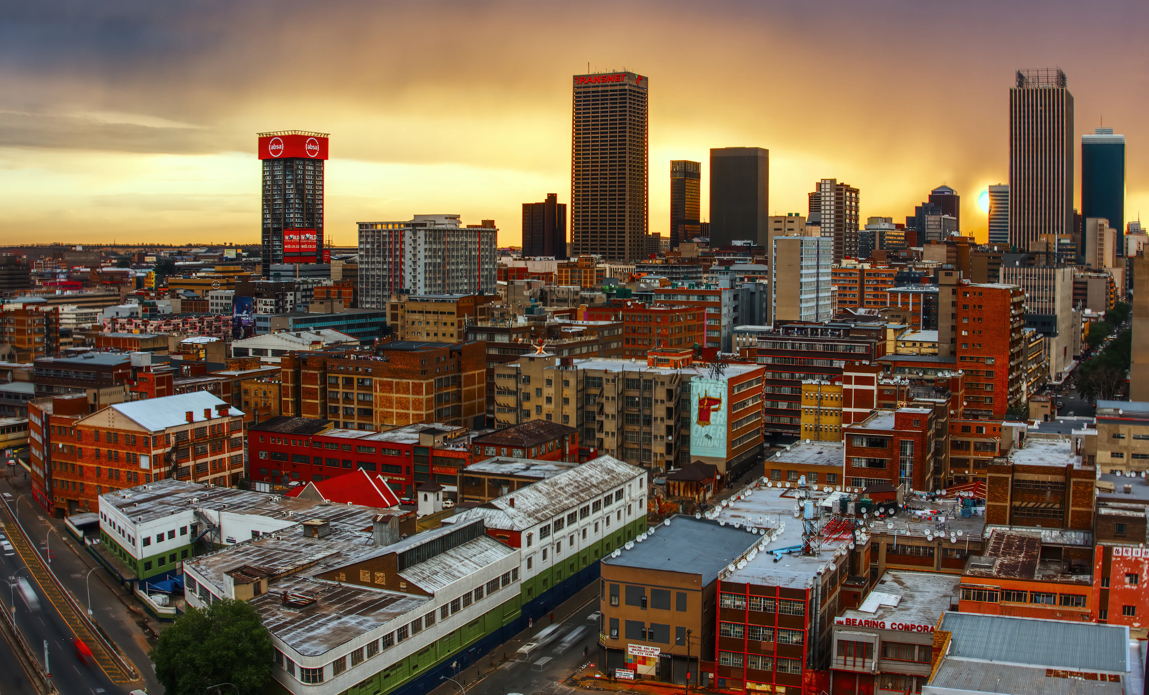 The inner city of Johannesburg at golden hour, its downtown towers standing dark against a burning orange sky above blocks of low warehouses, shopfronts and painted rooftops
