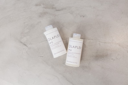 Two bottles of Olaplex hair care products are placed on a light gray surface. The bottles are labeled as No. 4 Bond Maintenance Shampoo and No. 5 Bond Maintenance Conditioner. The packaging is simple and minimalistic, showcasing white bottles with black text.