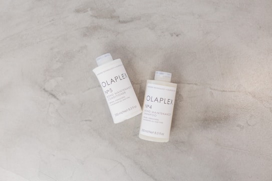 Two bottles of Olaplex hair care products are placed on a light gray surface. The bottles are labeled as No. 4 Bond Maintenance Shampoo and No. 5 Bond Maintenance Conditioner. The packaging is simple and minimalistic, showcasing white bottles with black text.