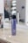 A bottle of MATRIX Total Results So Silver shampoo is placed on a wooden surface. The background features a mirror with reflections and blurred objects, including a person sitting and some salon equipment.