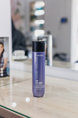 A bottle of MATRIX Total Results So Silver shampoo is placed on a wooden surface. The background features a mirror with reflections and blurred objects, including a person sitting and some salon equipment.