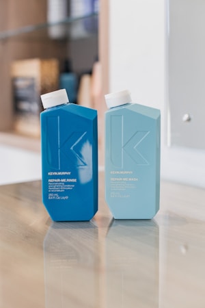 Two bottles of hair care products are placed on a reflective wooden surface. The left bottle is dark blue labeled 'REPAIR-ME.RINSE' and the right bottle is light blue labeled 'REPAIR-ME.WASH'. The background is softly blurred, suggesting a shelf with various items.