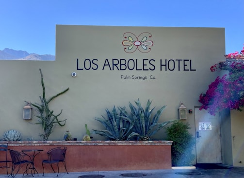 A hotel exterior wall with the name 'Los Arboles Hotel' and 'Palm Springs, Ca.' written on it. The wall is adorned with various desert plants, including large cacti and agave. Two metal chairs and a round table are in front of a low brick wall, and vibrant pink bougainvillea climbs one side of the wall. A light sconce is mounted beside the door on the wall.