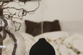 A minimalist bedroom setting with a focus on a black aroma diffuser emitting a thin stream of vapor. In the background, a glass vase holds a twisted, decorative branch, and a blurred bed with cream-colored bedding and a brown pillow can be seen.