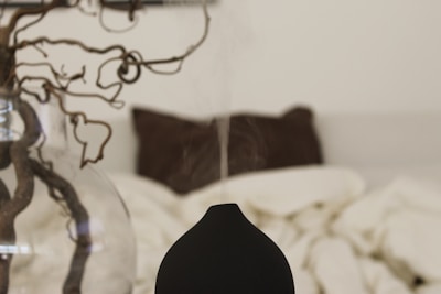 A minimalist bedroom setting with a focus on a black aroma diffuser emitting a thin stream of vapor. In the background, a glass vase holds a twisted, decorative branch, and a blurred bed with cream-colored bedding and a brown pillow can be seen.