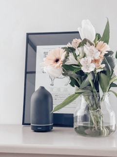 A bouquet of flowers in a glass vase is placed on a white surface next to a dark-colored essential oil diffuser. Behind the arrangement, there is a framed piece of artwork featuring sketches or illustrations.
