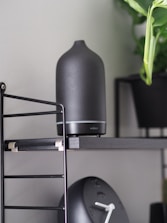 A matte black aroma diffuser is placed on a sleek black shelf with metal supports. Beside it, a black modern clock with white clock hands is visible. A green leafy plant in a black pot adds a touch of nature to the setting.