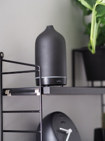 A matte black aroma diffuser is placed on a sleek black shelf with metal supports. Beside it, a black modern clock with white clock hands is visible. A green leafy plant in a black pot adds a touch of nature to the setting.