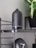 A matte black aroma diffuser is placed on a sleek black shelf with metal supports. Beside it, a black modern clock with white clock hands is visible. A green leafy plant in a black pot adds a touch of nature to the setting.
