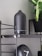 A matte black aroma diffuser is placed on a sleek black shelf with metal supports. Beside it, a black modern clock with white clock hands is visible. A green leafy plant in a black pot adds a touch of nature to the setting.