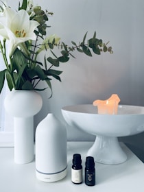 A serene setting with a white ceramic vase holding white lilies and green leaves on a white surface. Next to it is a sleek white essential oil diffuser, paired with two small bottles of essential oil. In the background, a white pedestal dish holds a small, softly glowing candle, contributing to a calming ambiance.