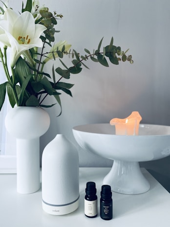 A serene setting with a white ceramic vase holding white lilies and green leaves on a white surface. Next to it is a sleek white essential oil diffuser, paired with two small bottles of essential oil. In the background, a white pedestal dish holds a small, softly glowing candle, contributing to a calming ambiance.