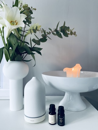 A serene setting with a white ceramic vase holding white lilies and green leaves on a white surface. Next to it is a sleek white essential oil diffuser, paired with two small bottles of essential oil. In the background, a white pedestal dish holds a small, softly glowing candle, contributing to a calming ambiance.
