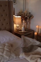 A cozy and elegantly decorated bedroom with a beige tufted headboard. A bedside table beside the bed holds a lit lamp with a beige lampshade, a candle, and a vase with dried flowers, creating a warm and inviting atmosphere. Various small bottles are also present on the table. Several lit candles are arranged on the floor beside the table, enhancing the serene and tranquil mood. A beige throw blanket and a decorative pillow with a textured design are laid on the bed.