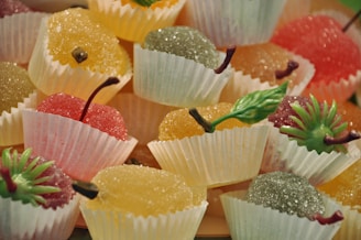 A collection of brightly colored, sugar-coated fruit jellies arranged in white paper cups. Each jelly is shaped like a small fruit and topped with a decorative plastic stem and leaf, adding to the realistic appearance. The fruits are presented in an assortment of colors including red, orange, yellow, and green, creating a vibrant and enticing display.