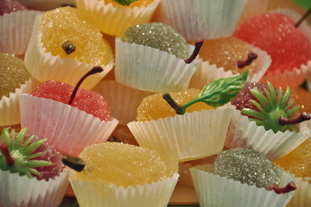 A collection of brightly colored, sugar-coated fruit jellies arranged in white paper cups. Each jelly is shaped like a small fruit and topped with a decorative plastic stem and leaf, adding to the realistic appearance. The fruits are presented in an assortment of colors including red, orange, yellow, and green, creating a vibrant and enticing display.