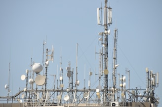 A variety of communication antennas and satellite dishes are mounted on a rooftop structure against a clear blue sky. The array includes different shapes and sizes of dishes, poles, and antennas, all made of metal and positioned at different heights.
