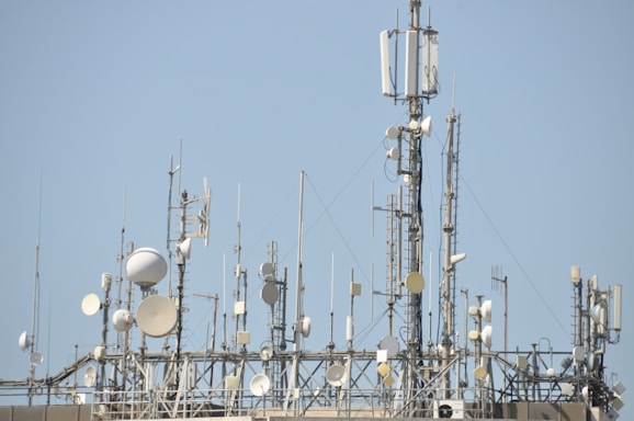 A variety of communication antennas and satellite dishes are mounted on a rooftop structure against a clear blue sky. The array includes different shapes and sizes of dishes, poles, and antennas, all made of metal and positioned at different heights.