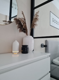 A minimalist room features a white dresser with decorative items on top, including a large white vase with dried grasses, a white candle holder, and a small black jar. A mirror reflects part of the room and is partially visible. A framed sign with the word 'GOOD' is mounted on the wall.