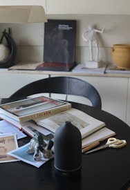 A tabletop filled with various art and design items, including an open book, magazines, and a small sculpture of rabbits. A black diffuser and a pair of scissors are also present. In the background, there is a shelf with books, a sculpture, and a ceramic pot.