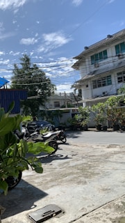 A street scene with several parked motorcycles on a concrete surface. There is a residential building in the background with visible windows and a slanted roof. Overhead, numerous power lines stretch across the sky. Lush green potted plants and trees line the surroundings, and the sky is partly cloudy with a clear blue hue.