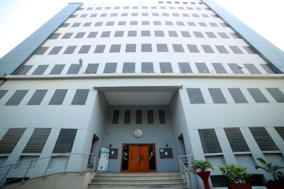 A tall, modern office building with a symmetrical facade and numerous rectangular windows. The entrance features a set of stairs leading to wooden double doors, flanked by potted plants. The building has a light color scheme with blue and gray tones, and the structure's clean lines and minimalistic design give it a contemporary feel.