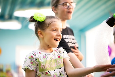 A young girl is smiling and looking with delight at bubbles coming from a machine. Her hair is styled with a bright green hair accessory, and she is wearing a shirt with a playful avocado pattern. Another person, slightly out of focus, stands beside her.