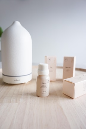 A minimalist arrangement of an essential oil diffuser and several small bottles and boxes of essential oils on a wooden surface. The diffuser is a smooth, white ceramic with a simple, elegant shape. The bottles and boxes are beige with soft, neutral tones, giving the setting a calm and soothing feel.