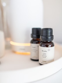 Two small brown glass bottles with black caps sit on a white tray. The bottles have labels indicating they are essential oils, one of which is named 'Breathe'. In the background, there is a white device emitting a soft, warm glow.