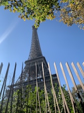 An upward view of a tall metal structure, framed by a fence in the foreground with lush green leaves and branches. The structure is set against a clear blue sky, partially obscured by leafy branches at the top.