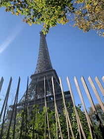 An upward view of a tall metal structure, framed by a fence in the foreground with lush green leaves and branches. The structure is set against a clear blue sky, partially obscured by leafy branches at the top.