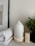 A serene setup featuring a white essential oil diffuser with a warm light at its base, placed on a smooth surface. Beside it is a neatly rolled, soft white towel and a small box labeled 'Relax' containing essential oils. A small potted plant with green leaves adds a touch of nature to the scene.