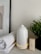 A serene setup featuring a white essential oil diffuser with a warm light at its base, placed on a smooth surface. Beside it is a neatly rolled, soft white towel and a small box labeled 'Relax' containing essential oils. A small potted plant with green leaves adds a touch of nature to the scene.