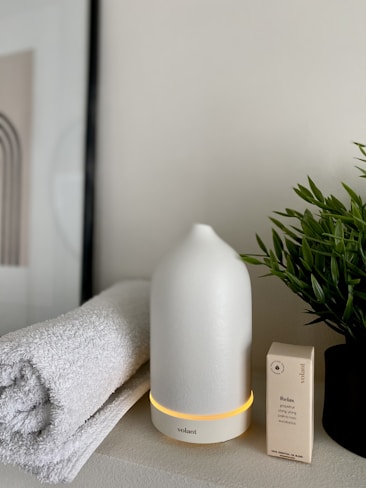 A serene setup featuring a white essential oil diffuser with a warm light at its base, placed on a smooth surface. Beside it is a neatly rolled, soft white towel and a small box labeled 'Relax' containing essential oils. A small potted plant with green leaves adds a touch of nature to the scene.